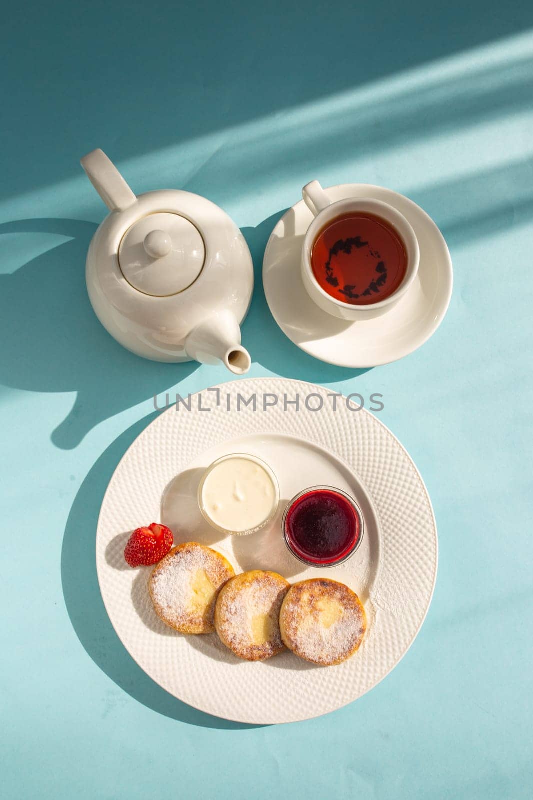 White plate with cottage cheese pancakes, sour cream, jam, and a cup of tea on a blue table. by Pukhovskiy