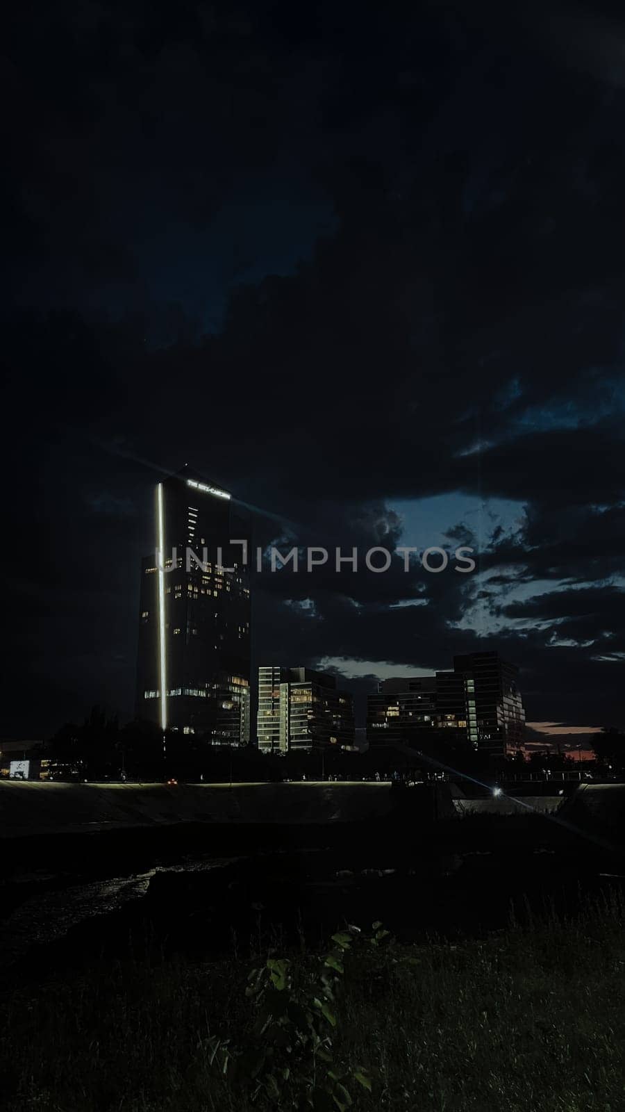 A tall skyscraper stands out among other buildings in the city at night. The sky is dark and cloudy, with a hint of light on the horizon.