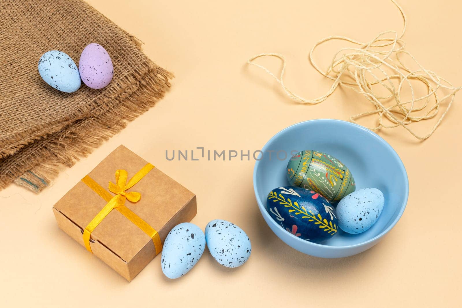 Blue bowl with colored Easter eggs, a sackcloth bag with eggs, a gift box and a rope. Top view.