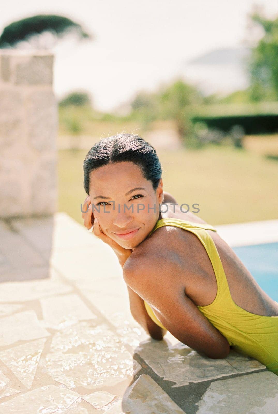 Smiling young woman resting her chin on her hand while standing in the pool and leaning on the side. High quality photo