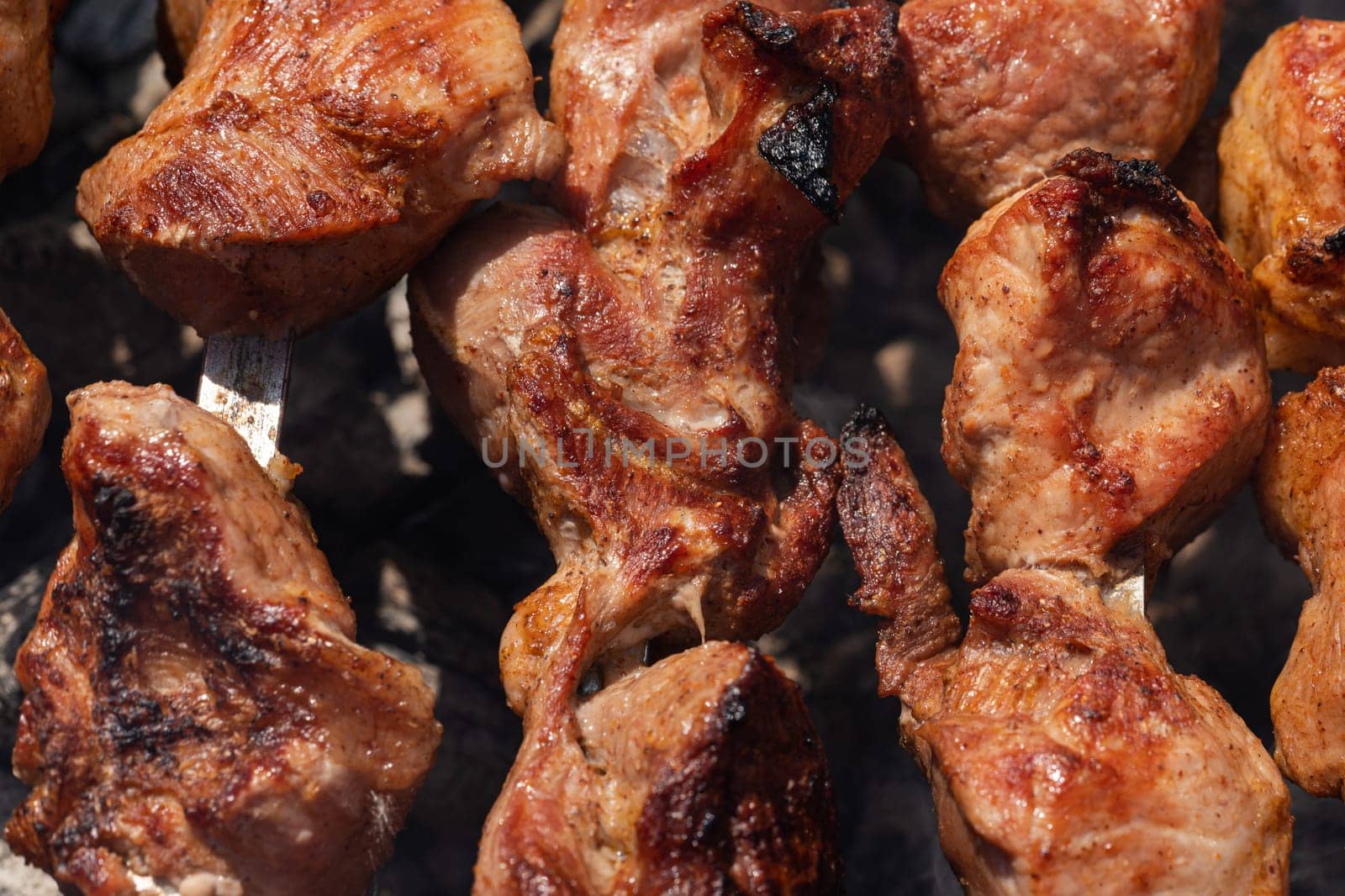 Cooking during summer picnic - appetizing juicy pork shish kebabs on metal skewers on charcoal grill with fragrant fire smoke. Close-up view, selective focus on tasty pieces of roast meat.