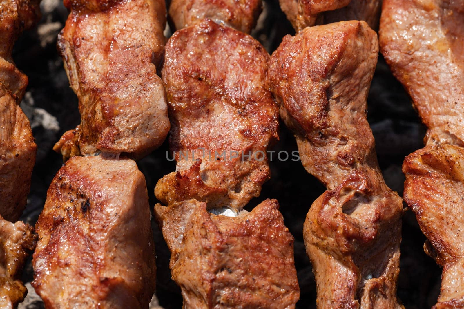 Appetizing juicy pork shish kebabs cooking on metal skewers on charcoal grill with fragrant smoke. Close-up view, selective focus on tasty pieces of roast meat. Cooking during summer picnic.
