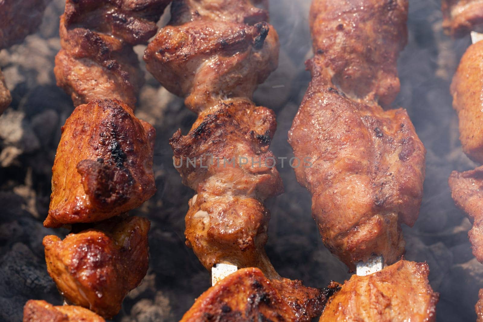 Grilled appetizing pork shish kebab cooking on metal skewers on charcoal grill with fragrant fire smoke. Cooking during summer picnic. Close-up view, selective focus on pieces of tasty roast meat.