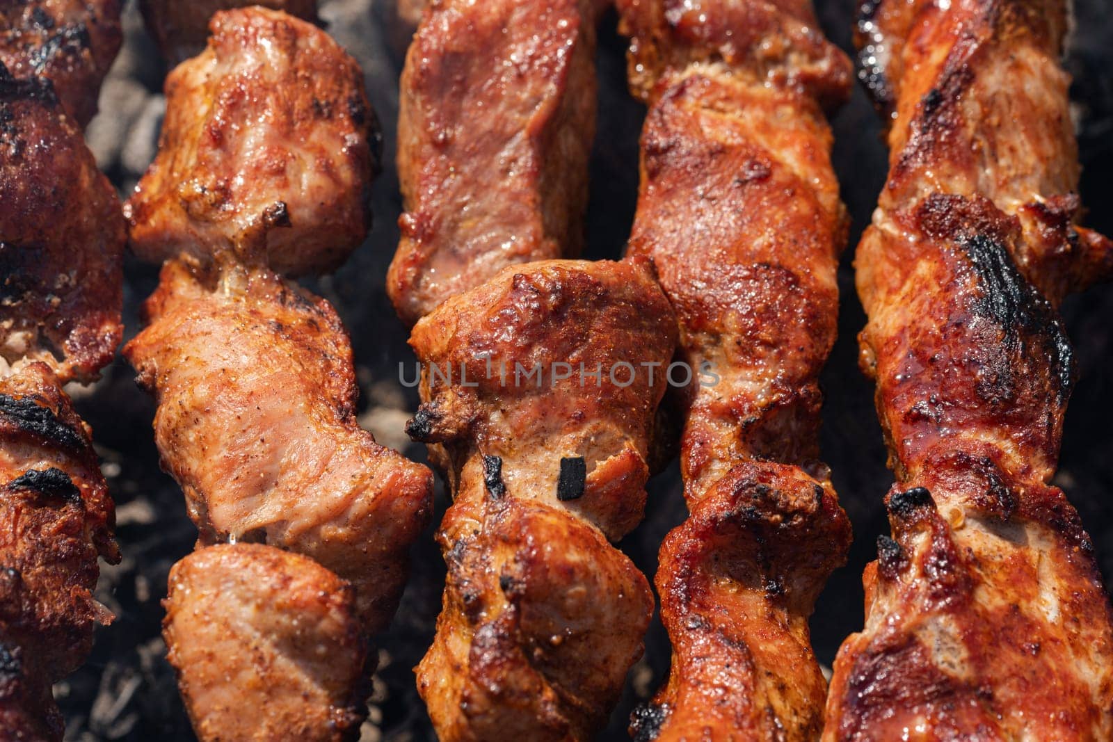 Tasty juicy pork barbecue cooking on metal skewers on outdoors charcoal grill with fragrant fire smoke. Close-up view, cooking during summer picnic. Selective focus on pieces of delicious roast meat.