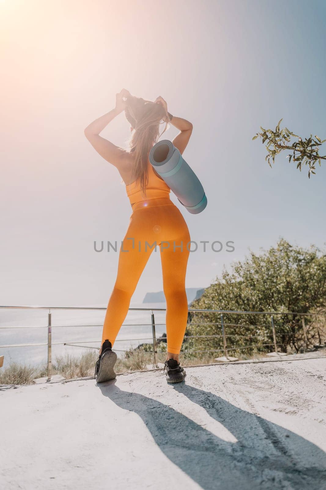 Fitness woman sea. Happy middle aged woman in orange sportswear exercises morning outdoors on yoga mat with laptop in park over ocean beach. Female fitness pilates yoga routine. Healthy lifestyle. by panophotograph