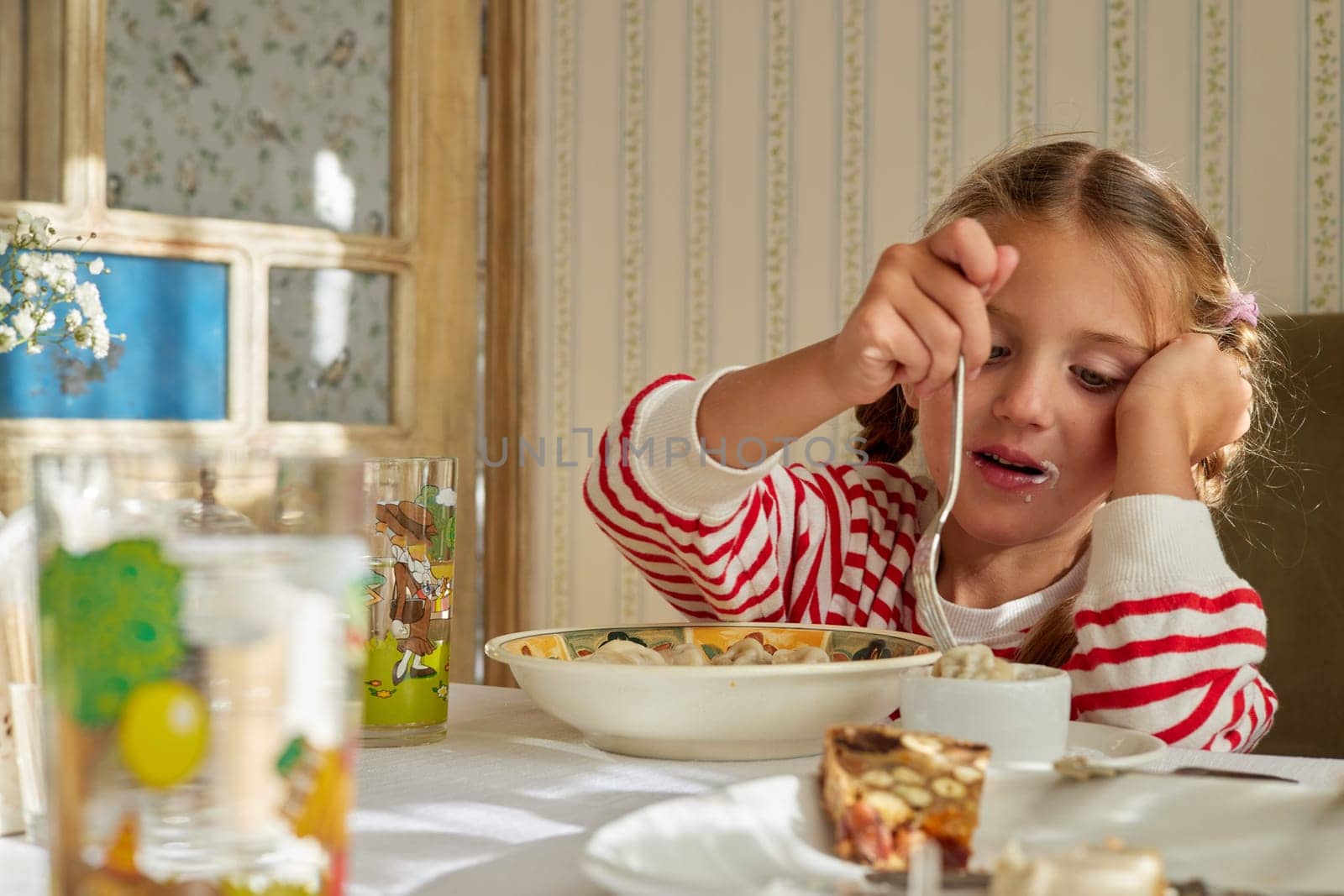 Adorable little girl with dirty mouth wearing red striped sweater sitting at table leaning on hand while dipping dumplings into sour cream with fork during lunch in kitchen with sunlight