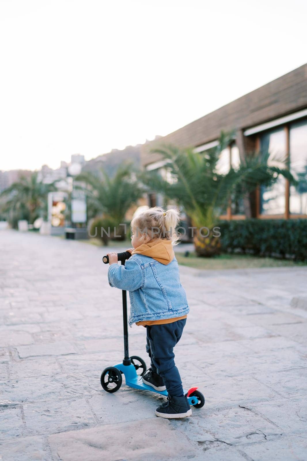 Little girl rides a scooter on the road past houses. Back view. High quality photo