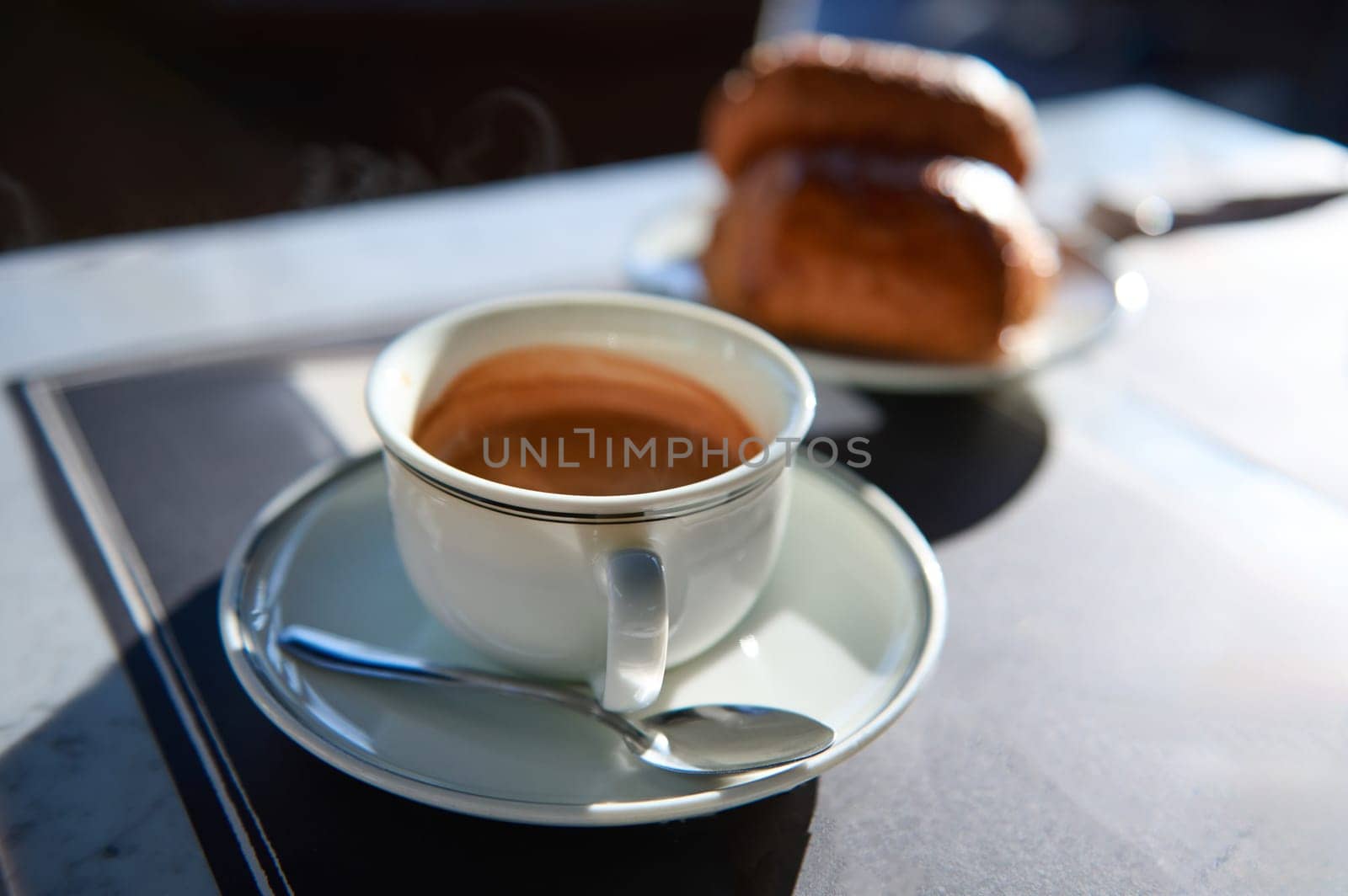 Elegant white ceramic cup of freshly brewed espresso coffee with foam on saucer. Blurred crispy croissants on background by artgf