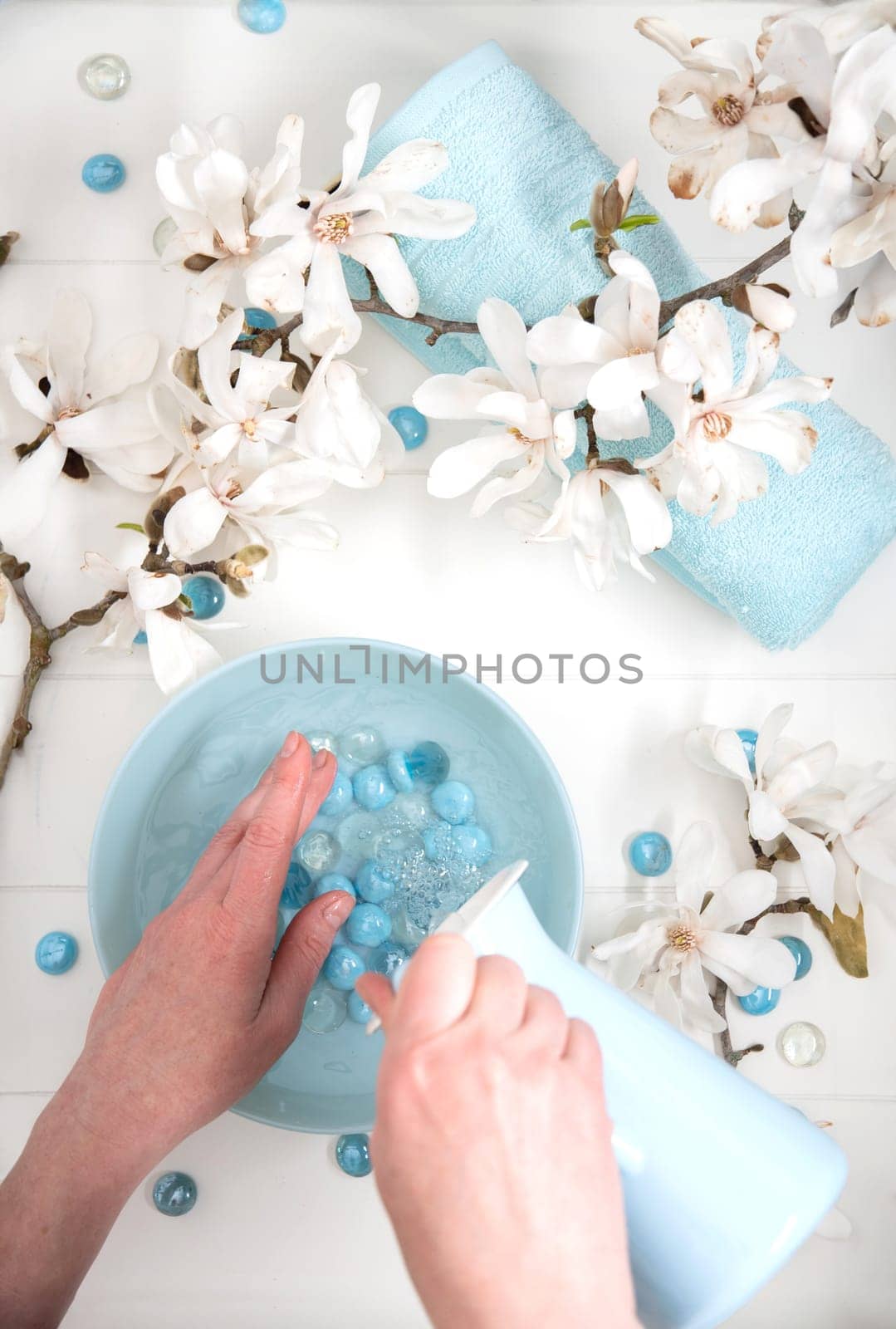 woman pours spring water into a bowl and sea pebbles for a delicate spa manicure procedure in a salon with white magnolia flowers on the table,High quality photo