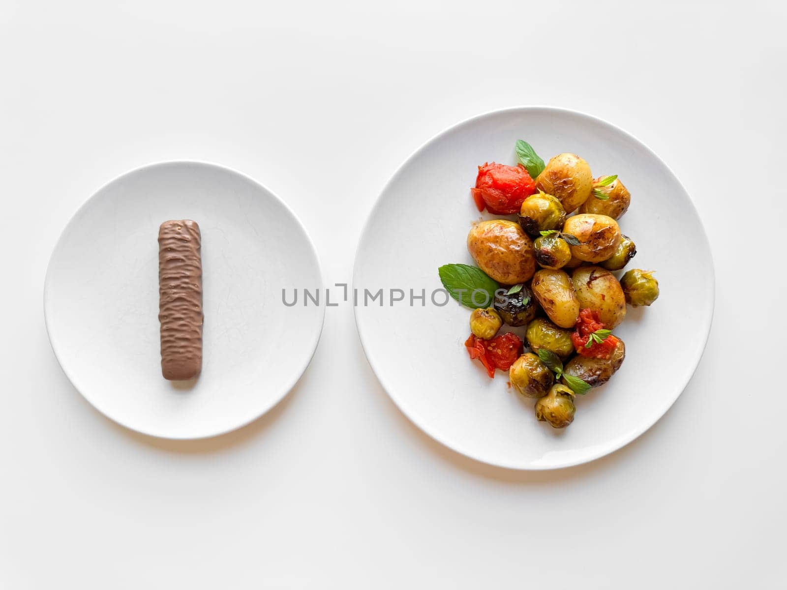 Two white plates on table, one with single chocolate bar and other with roasted baby potatoes and Brussels sprouts, contrasting healthy and indulgent food choices. by Lunnica