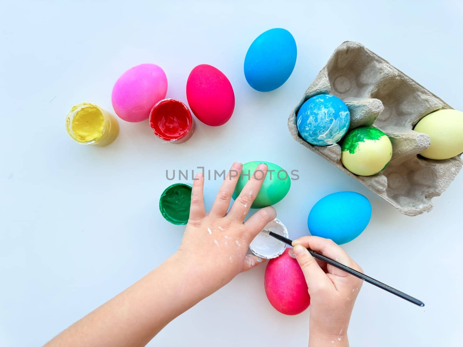 Childrens hands paint Easter eggs with brush surrounded by colorful eggs and jars of paints on white table, creative holiday activity. by Lunnica