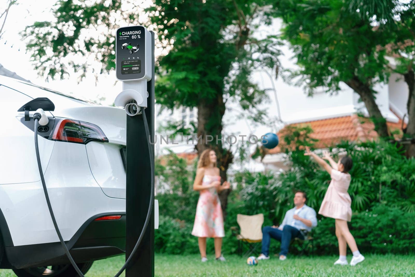 Focus electric vehicle recharge from home charging station on blur background of happy and playful family playing together. EV car using alternative and sustainable energy for better future.Synchronos