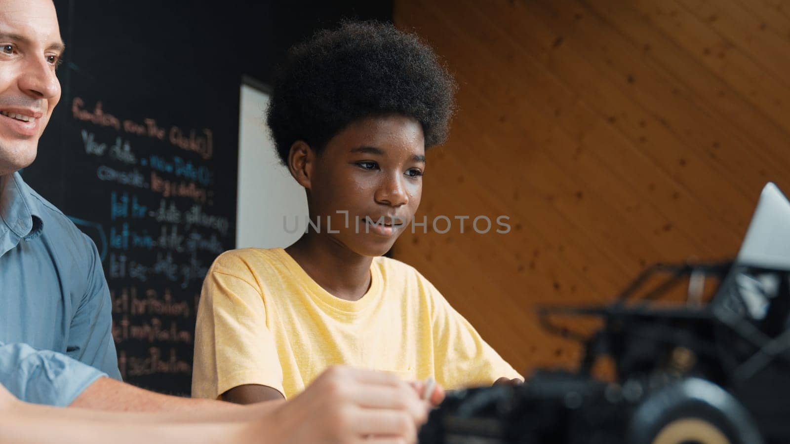 African boy using laptop to program code or prompt with car model placed while teacher and caucasian friend study about robotic construction. Student study about car model structure. Edification.