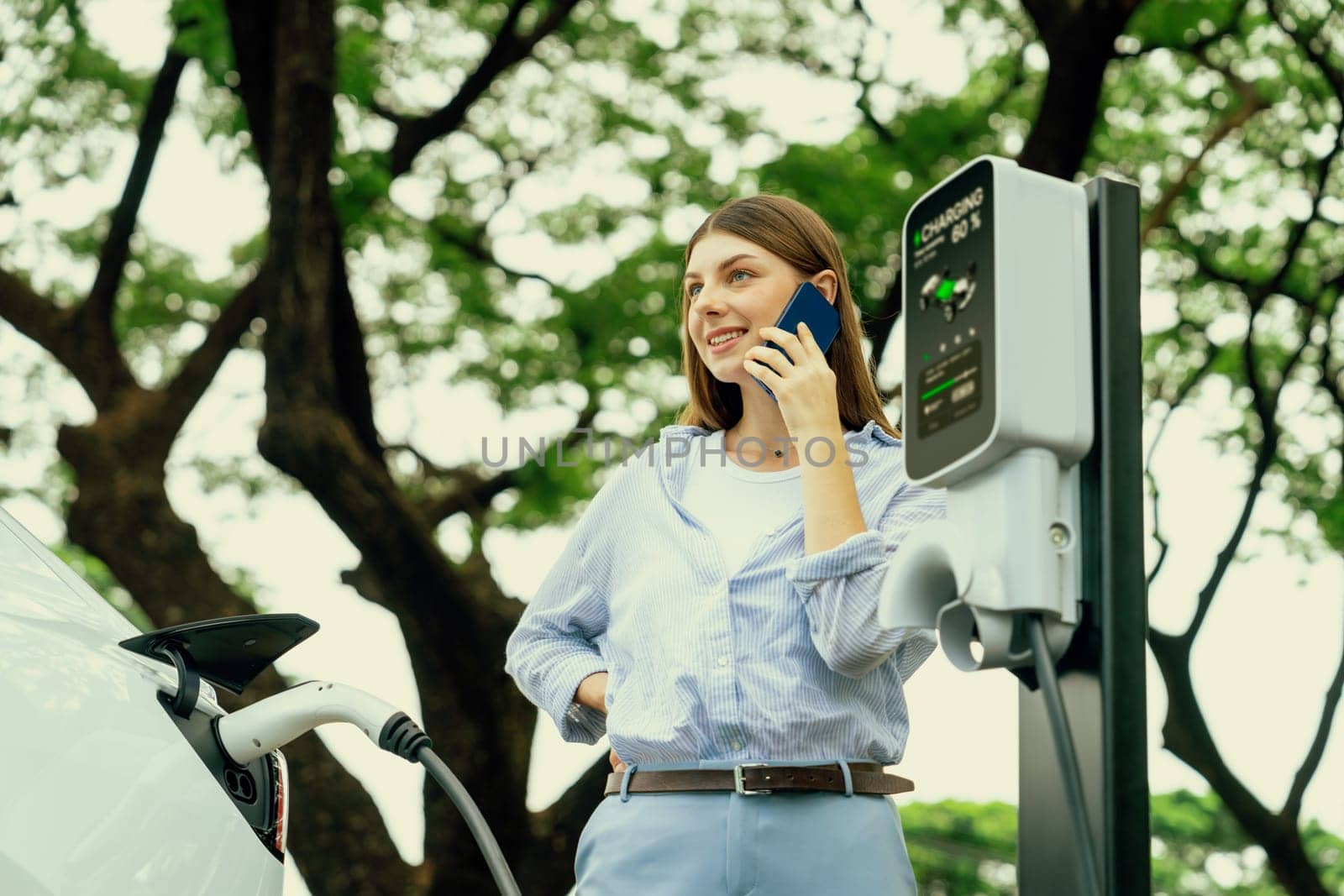 Young woman talking on smartphone while recharging electric car battery charging from EV charging station during vacation holiday road trip at national park or summer forest. Exalt
