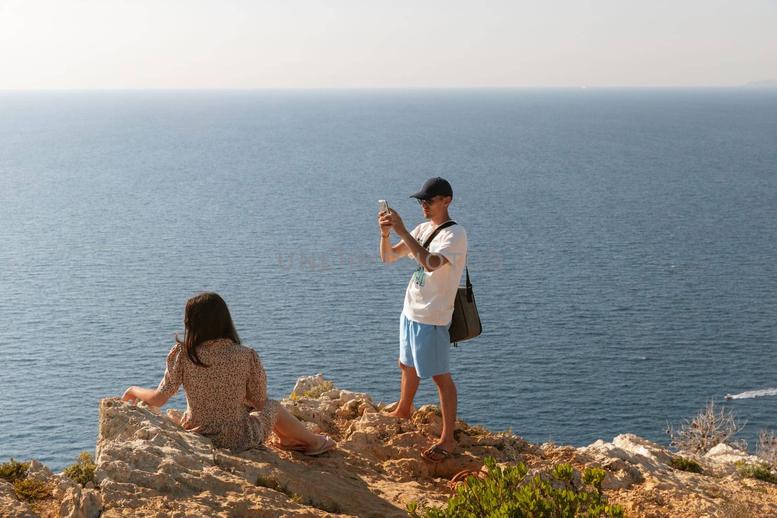 A young guy takes a photo of a girl on his phone by the sea. by Nataliya