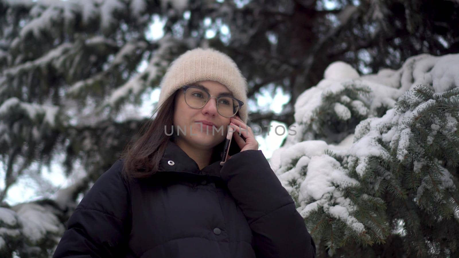 A young woman speaks on the phone near a tall spruce tree in winter. A girl with glasses communicates via cell phone against the backdrop of snowy spruce branches. by Puzankov