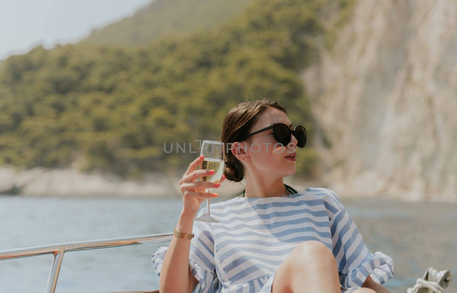 Portrait of one young Caucasian beautiful girl in sunglasses sitting in a boat, holding a glass of champagne in her hand, looking to the side and enjoying sailing on the sea on a private boat on a sunny summer day, close-up side view.