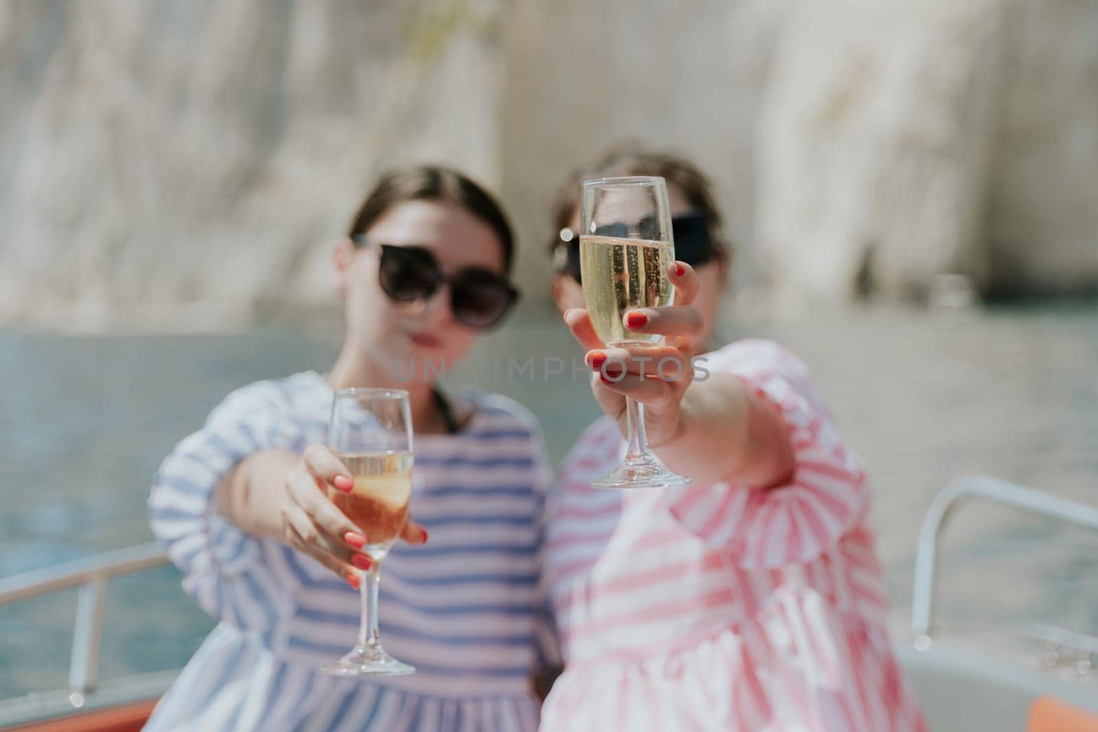 Portrait of one young Caucasian beautiful couple of girls in sunglasses sitting in a boat holding glasses of champagne forward against a background of blurred rocks while sailing on the sea on a sunny summer day, side view close-up with selective focus.