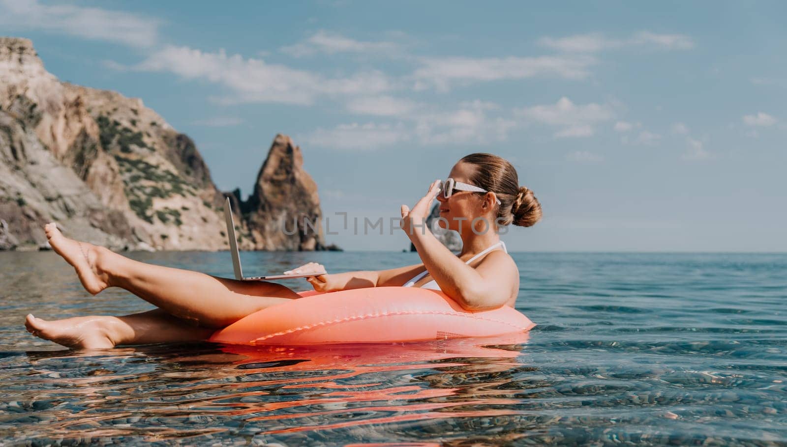 Woman freelancer works on laptop swimming in sea on pink inflatable ring. Happy tourist in sunglasses floating on inflatable donut and working on laptop computer in calm ocean. Remote working anywhere by panophotograph