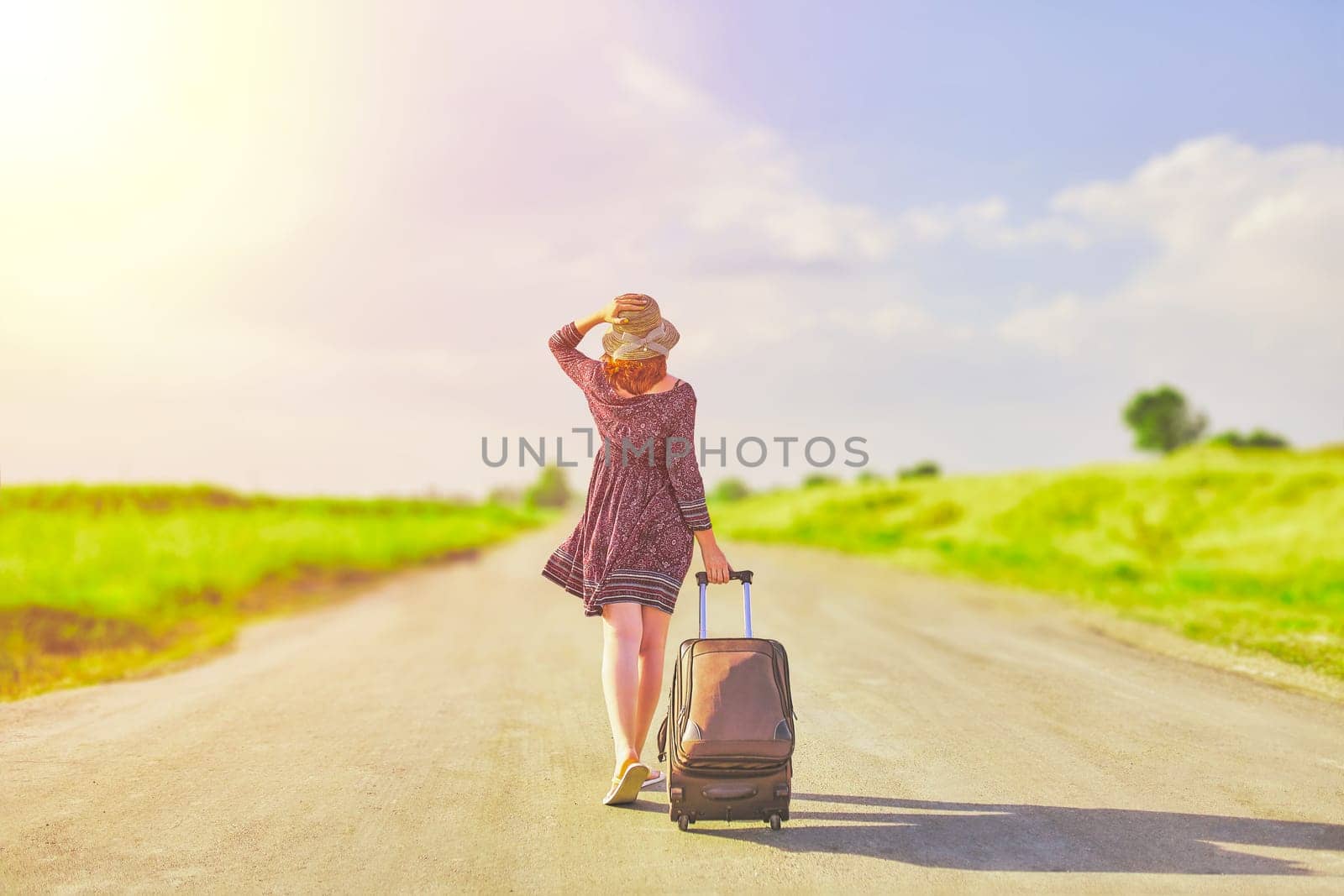 Beautiful woman in summer dress and hat hitchhiking with suitcase at spring. Female walking on the road with baggage at sunny day. Hitchhiker concept