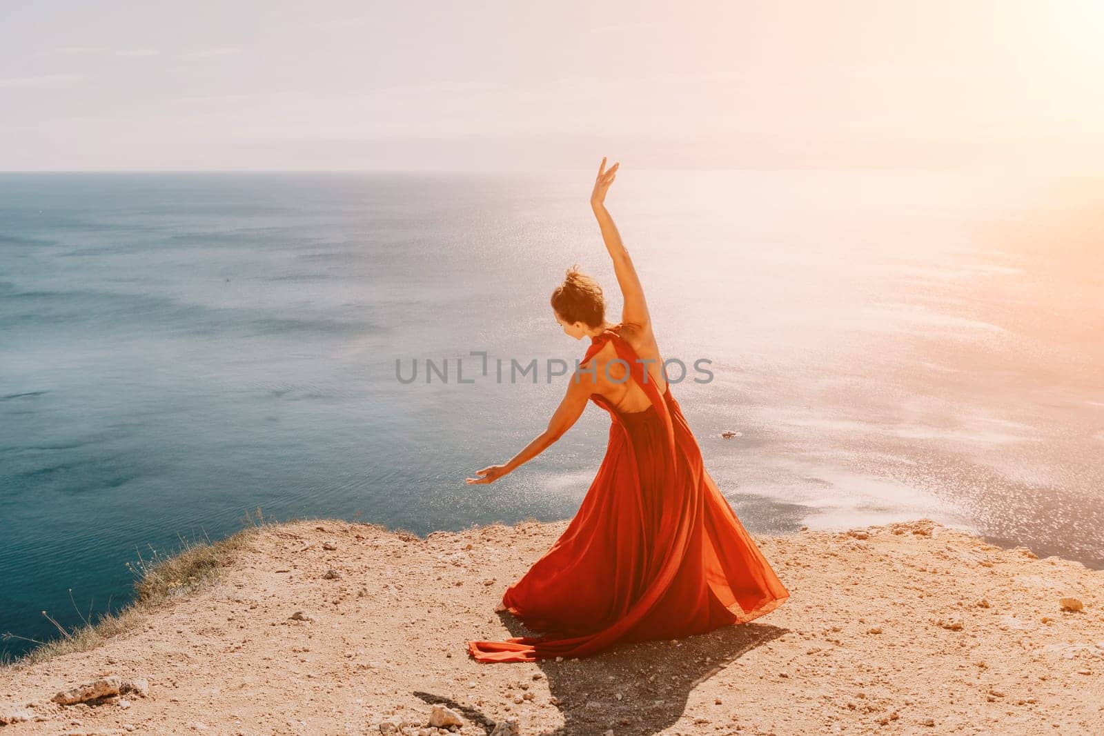 Side view a Young beautiful sensual woman in a red long dress posing on a rock high above the sea during sunrise. Girl on the nature on blue sky background. Fashion photo.