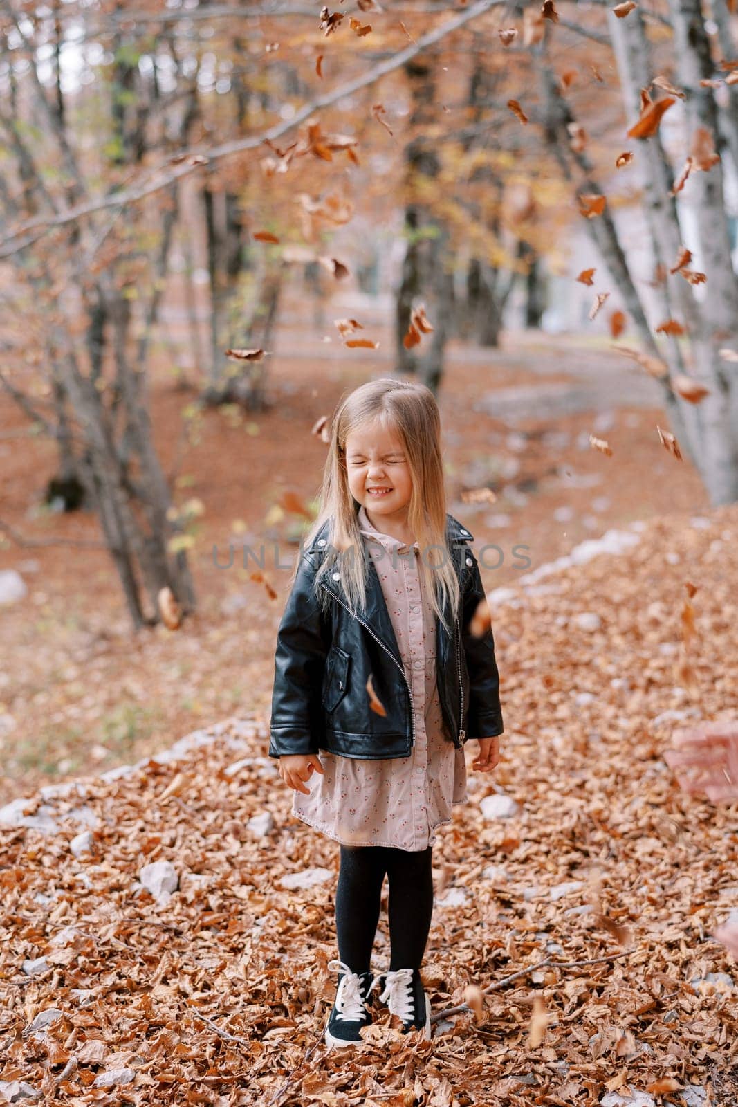 Little girl stands with her eyes closed under falling dry leaves in the autumn forest. High quality photo