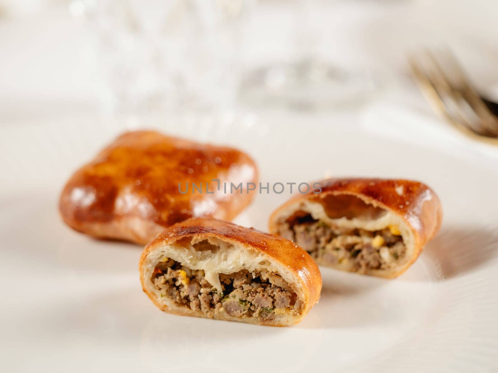 Small russian pies with minced meat Russian Piroshki. Cooked meat hand portioned pies or empanadas cut in half on white plate in elegant restaurant interior. Selective focus, close up.