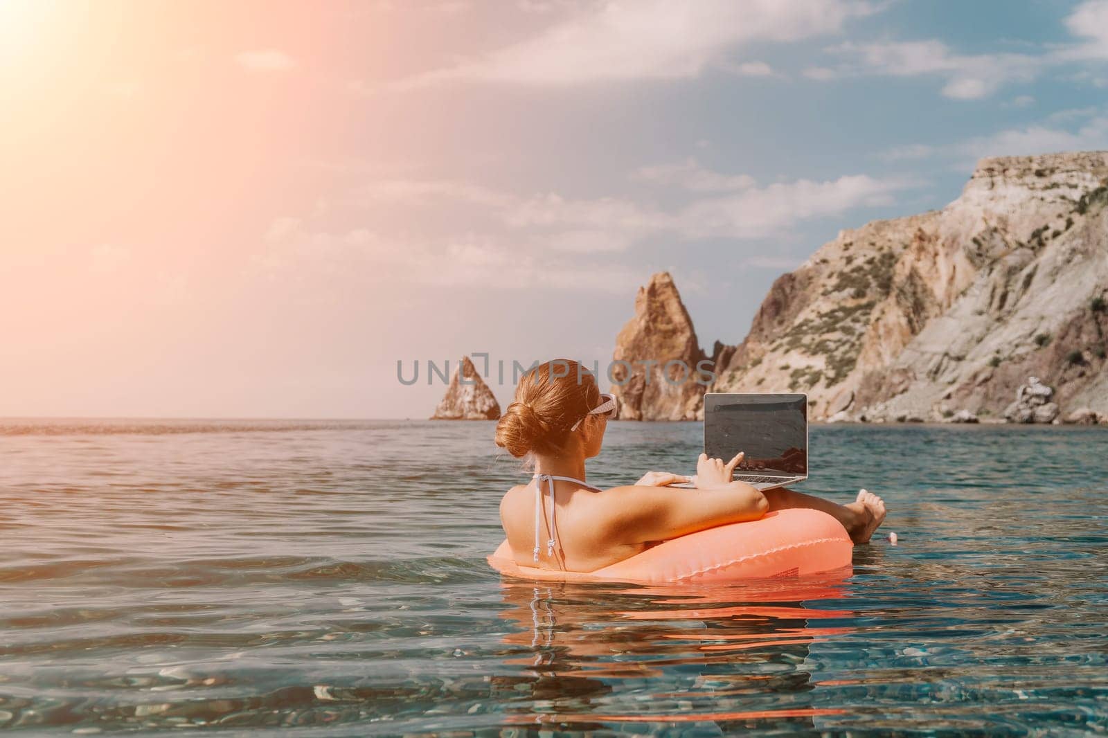 Woman freelancer works on laptop swimming in sea on pink inflatable ring. Happy tourist in sunglasses floating on inflatable donut and working on laptop computer in calm ocean. Remote working anywhere by panophotograph