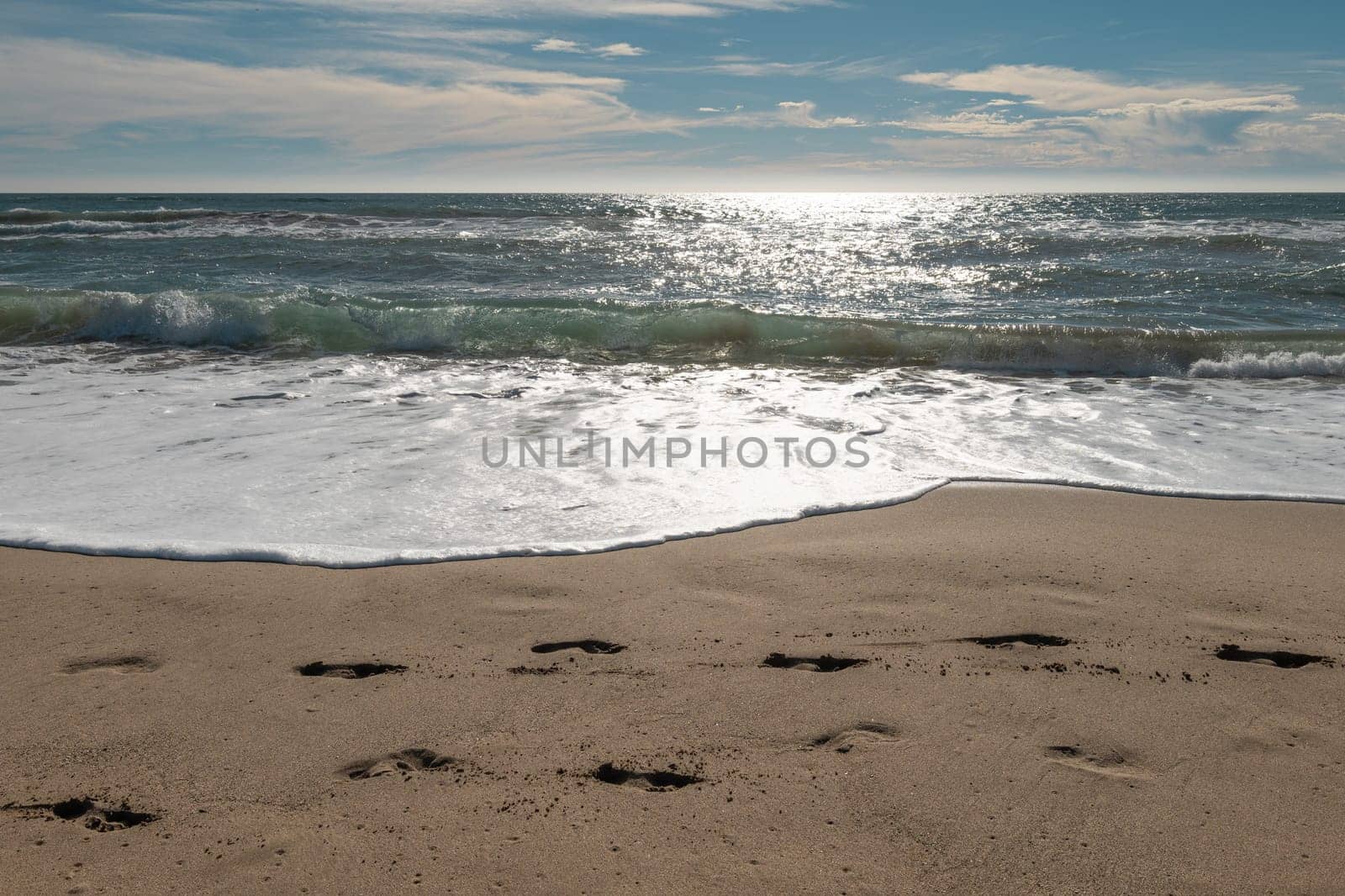 Footprints and Frothy Ocean Waves on Sandy Beach with Bright Sunlight Reflecting on Water by apavlin