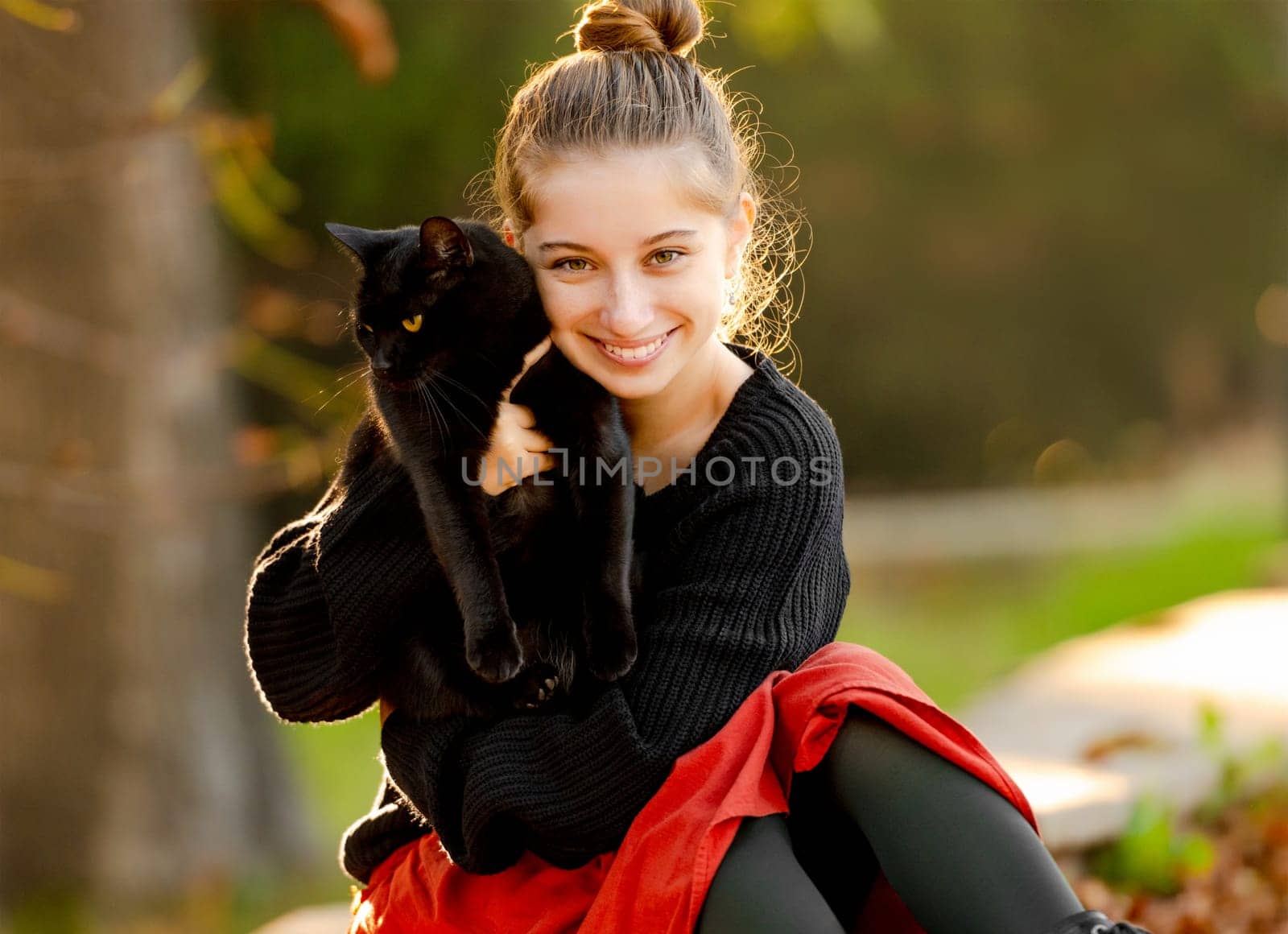 Pretty girl hugging black cat outdoors at street with autumn leaves portrait. Beautiful model teenager with feline animal at park in fall season