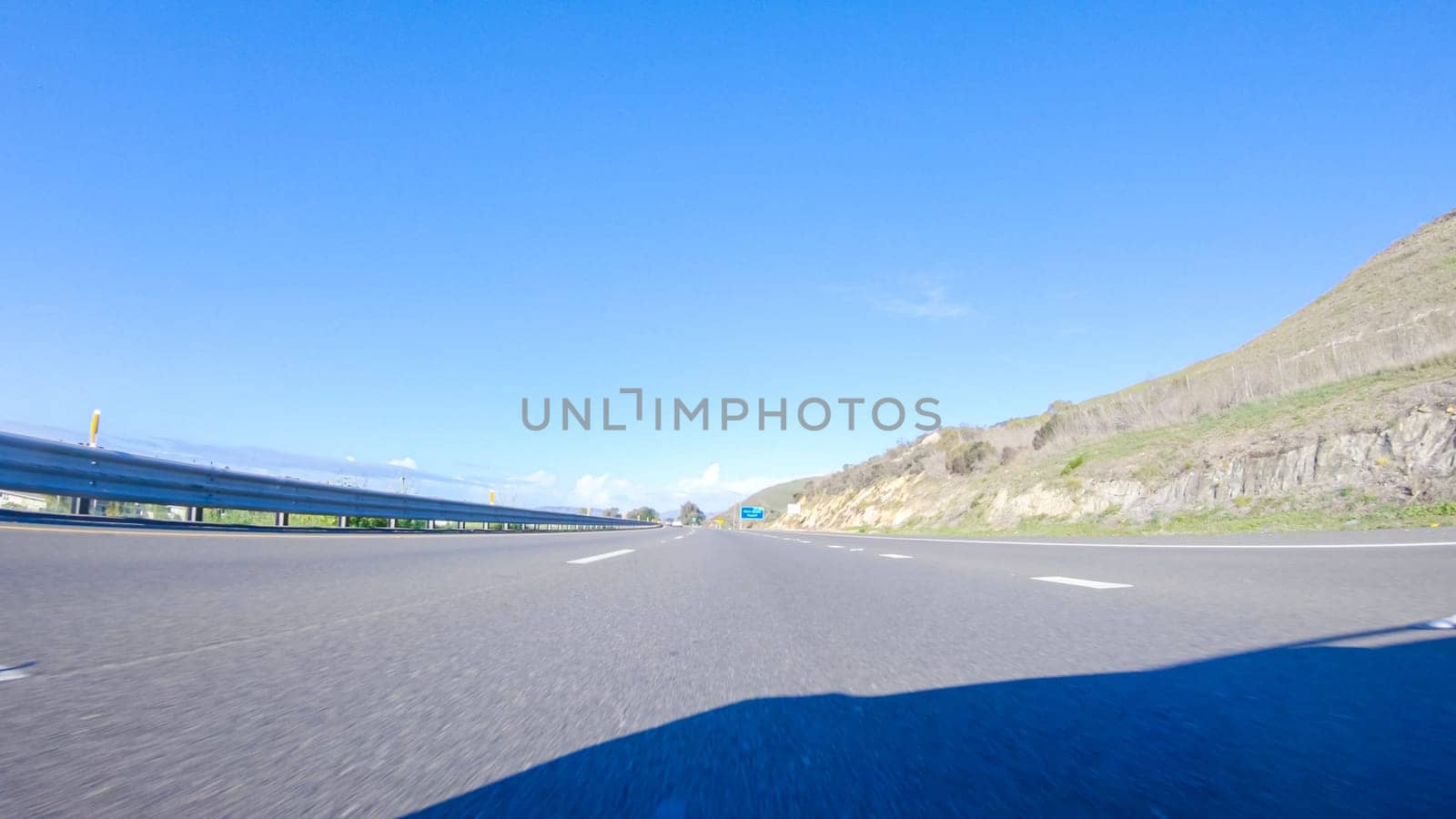 On a crisp winter day, a car cruises along the iconic Highway 1 near San Luis Obispo, California. The surrounding landscape is brownish and subdued, with rolling hills and patches of coastal vegetation flanking the winding road.