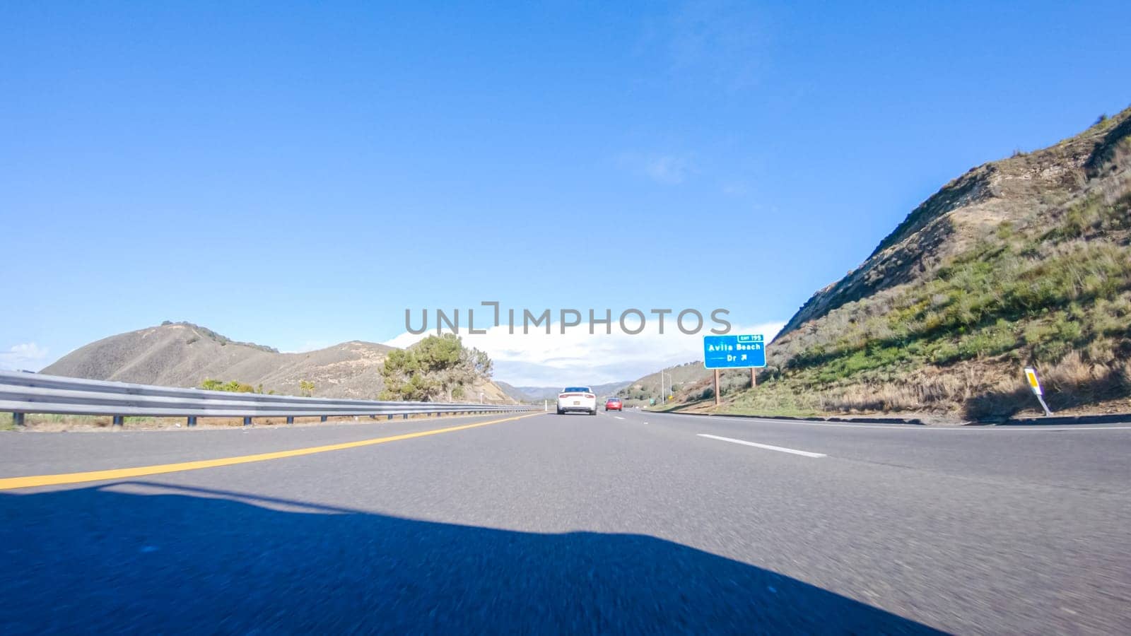 On a crisp winter day, a car cruises along the iconic Highway 1 near San Luis Obispo, California. The surrounding landscape is brownish and subdued, with rolling hills and patches of coastal vegetation flanking the winding road.