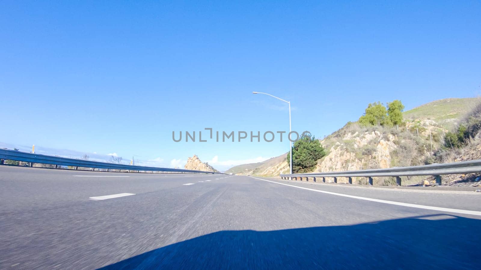 On a crisp winter day, a car cruises along the iconic Highway 1 near San Luis Obispo, California. The surrounding landscape is brownish and subdued, with rolling hills and patches of coastal vegetation flanking the winding road.