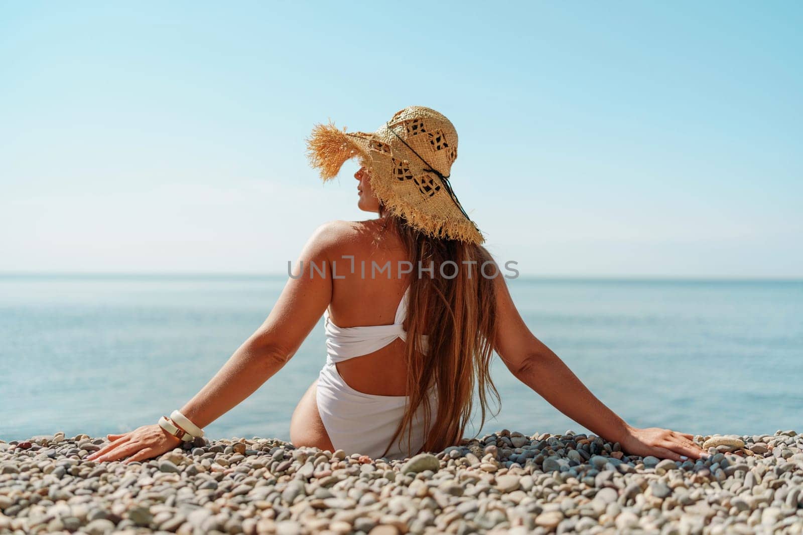 Beach Relaxation woman in hat sits on a pebble beach enjoying the sunshine. The concept of travel, vacation at sea.