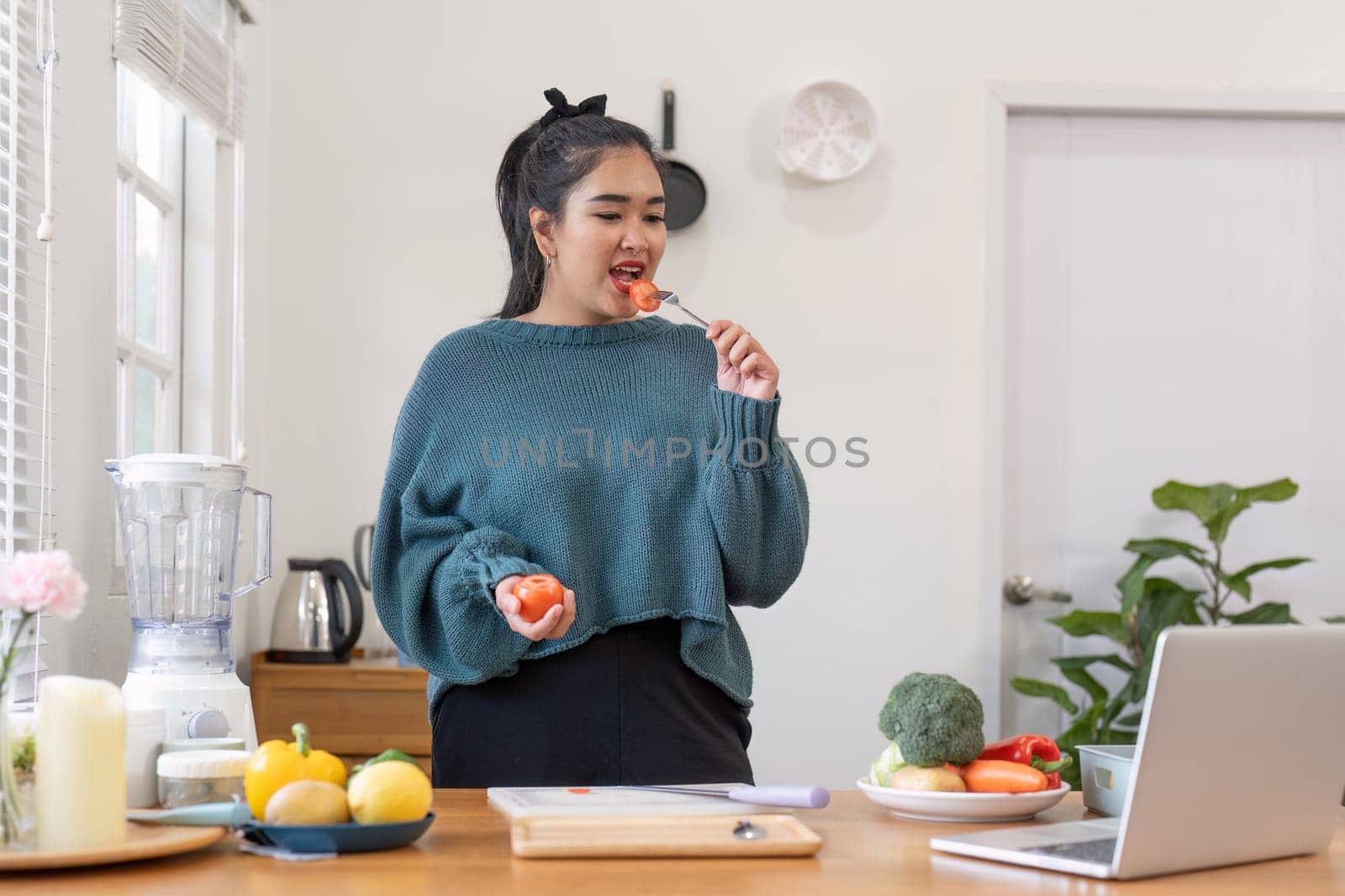 She is sitting at a table by the window, eating a tomato in front of her laptop. The laptop sleeve is made of wood, and there is house plant on the table