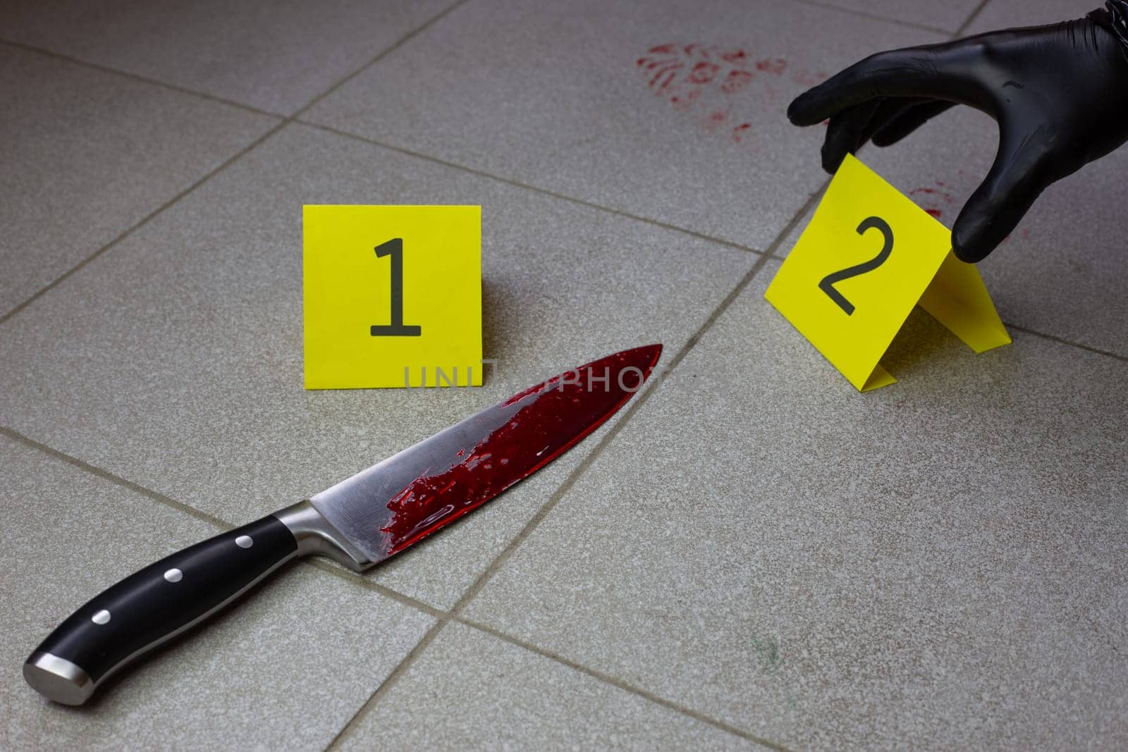 Criminologist in black gloves places sign with the evidence number near bloody knife at crime scene
