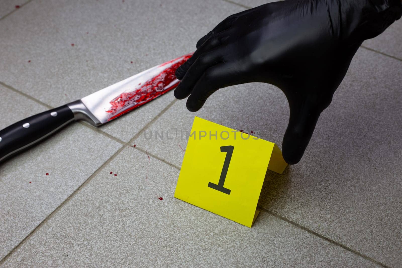 Criminologist in black gloves places yellow sign with the number of evidence at crime scene near bloody knife on the floor
