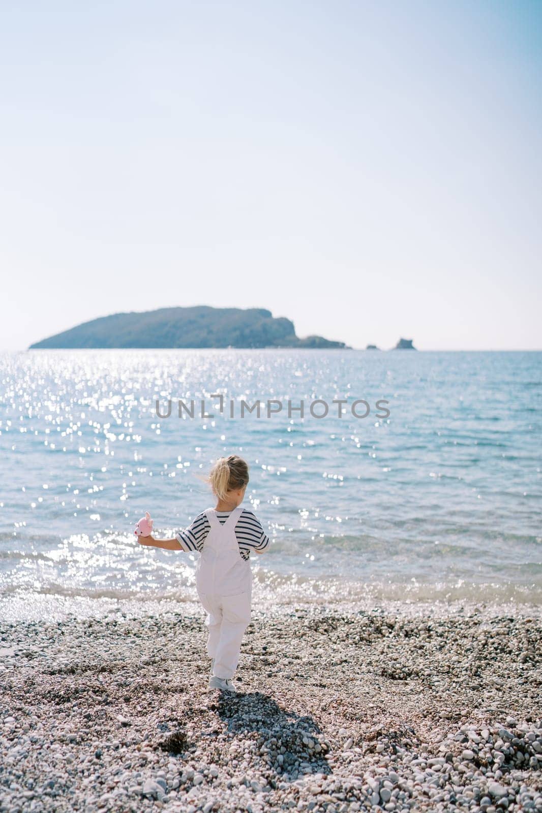 Little girl with a toy walks along a pebble beach towards the sea. Back view. High quality photo