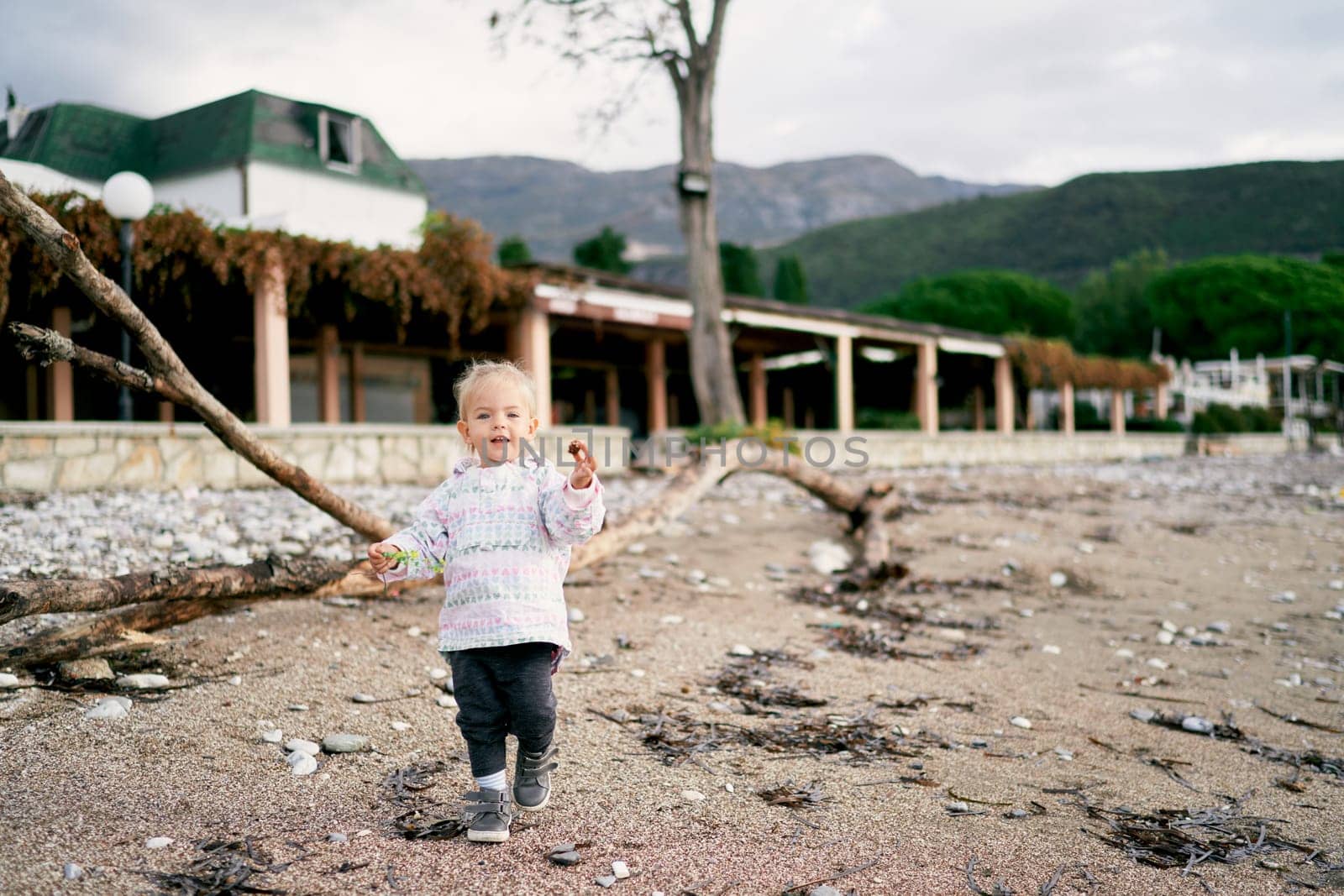 Little girl with a pebble in her hand walks along the beach against the backdrop of the house by Nadtochiy
