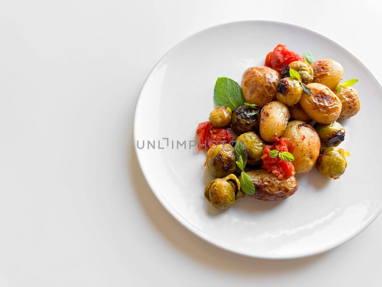 Roasted baby potatoes and Brussels sprouts with cherry tomatoes on white plate, garnished with basil, healthy vegetarian dish concept with space for text. High quality photo