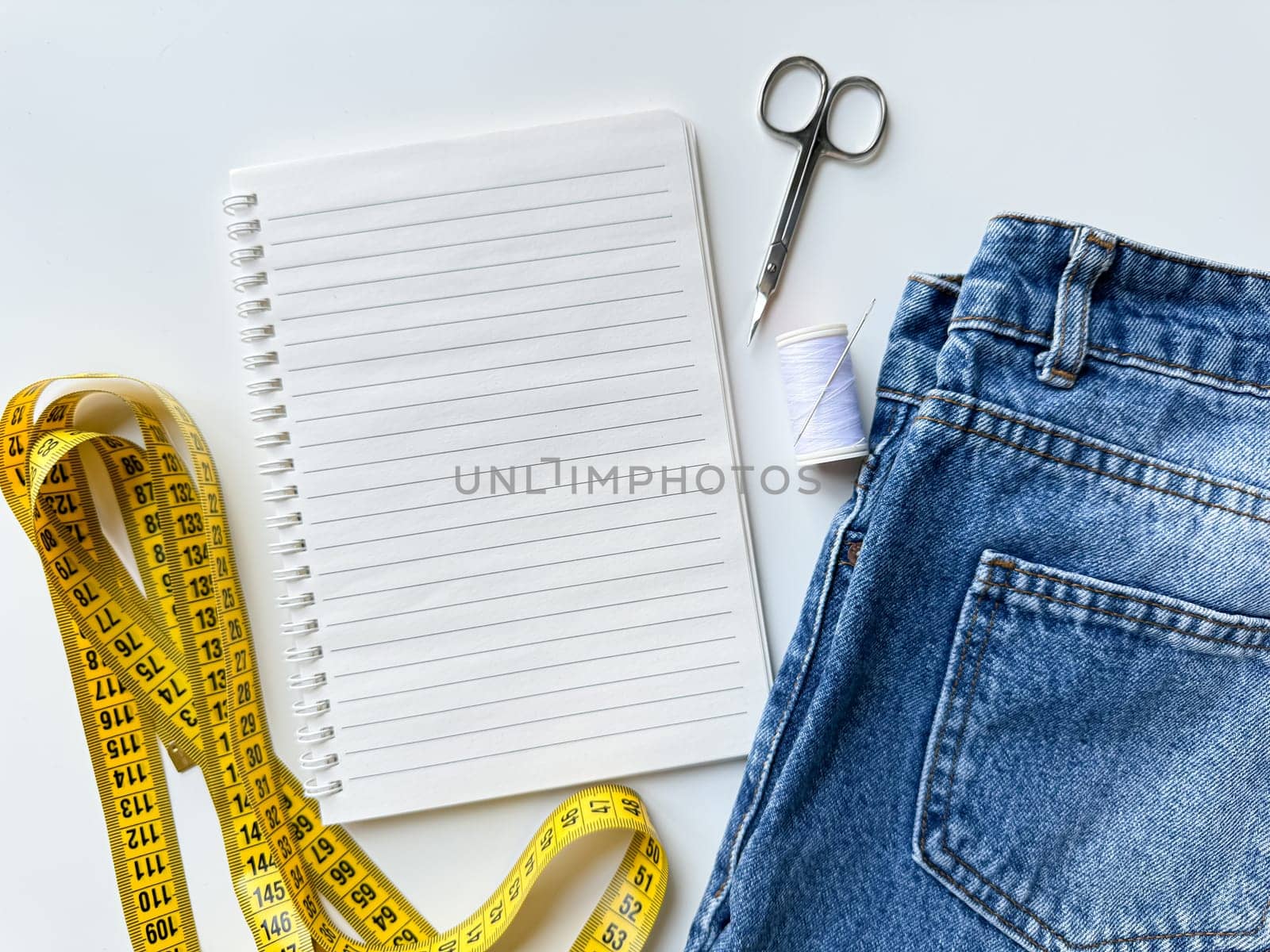 Tailoring and fashion design concept with denim jeans, yellow measuring tape, scissors, thread, and notepad with blank page for text on white background for planning and alterations. Top view. by Lunnica