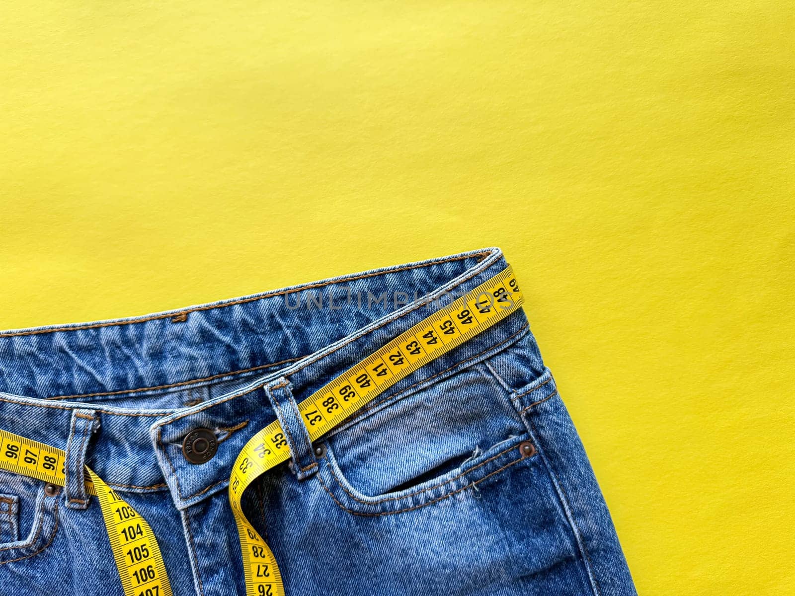 Close up of blue denim jeans with yellow measuring tape belt on vivid yellow background with copy space. Fashion, dieting, and body measurements. Top view. High quality photo