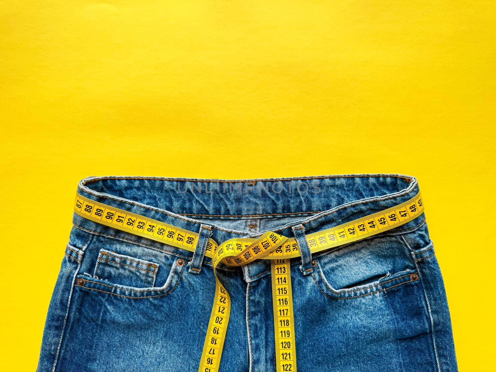 Blue jeans with measuring tape as belt on bright yellow background with copy space. Depicting weight loss, diet, and healthy lifestyle concept. Top view. High quality photo