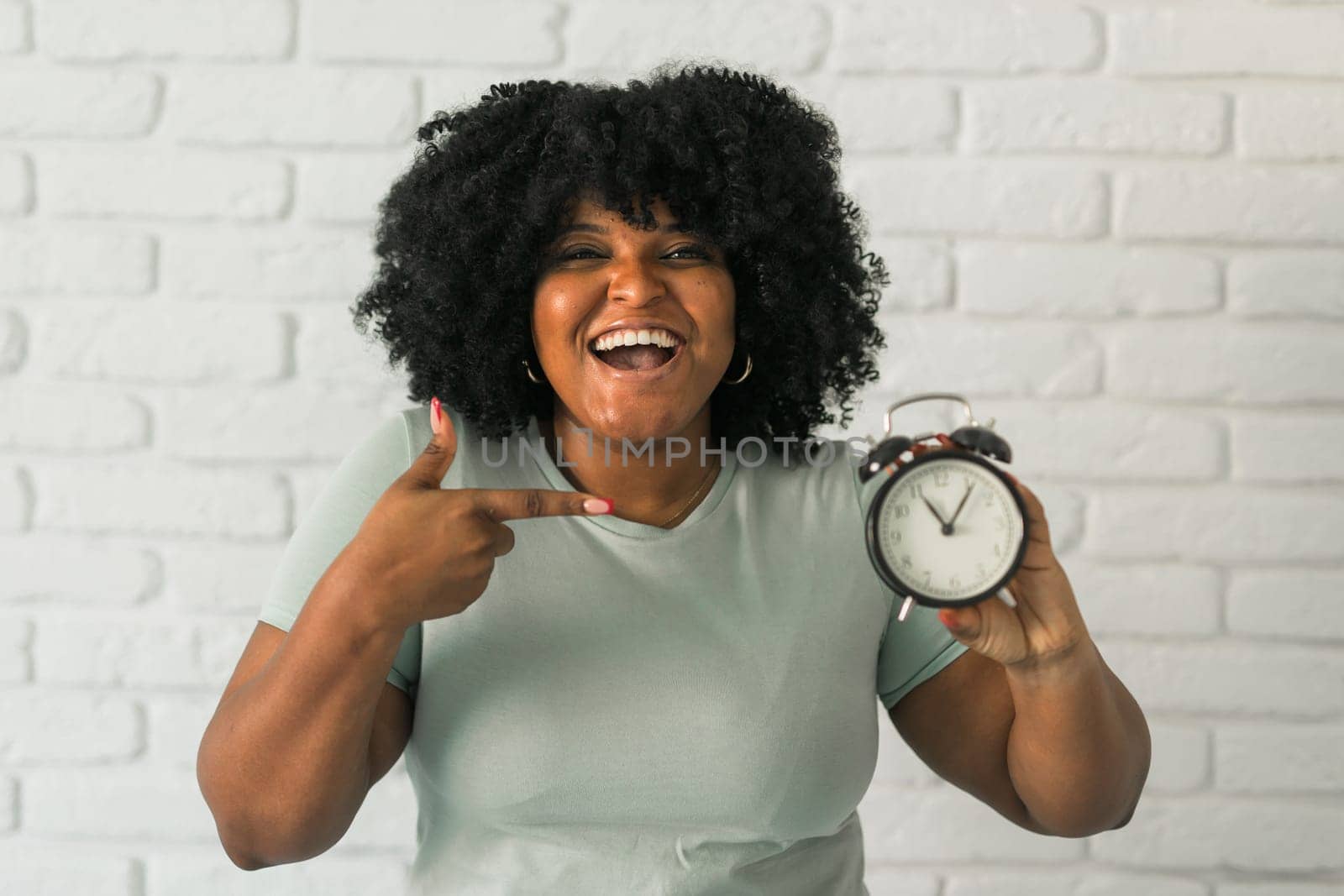Surprised african american woman holding alarm clock amazed with open mouth and surprise happy face at home, on brick background. Being late and time passing quickly concept by Satura86