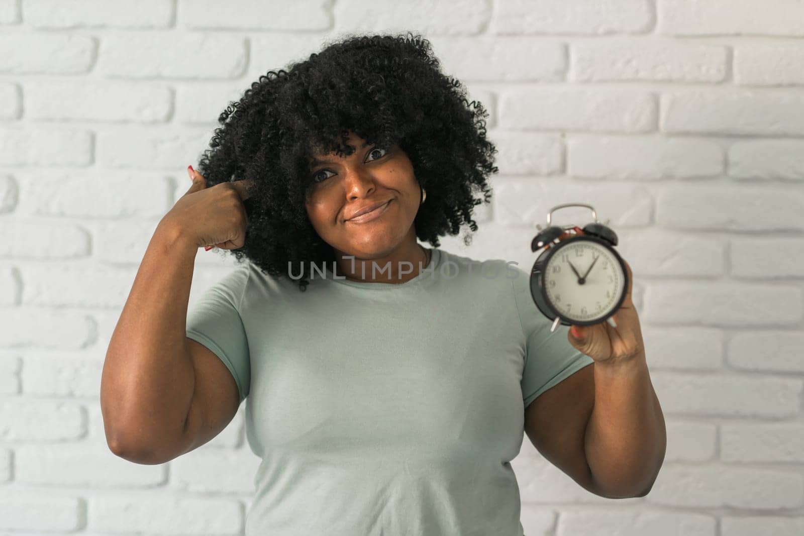 Surprised african american woman holding alarm clock amazed with open mouth and surprise happy face at home, on brick background. Being late and time passing quickly