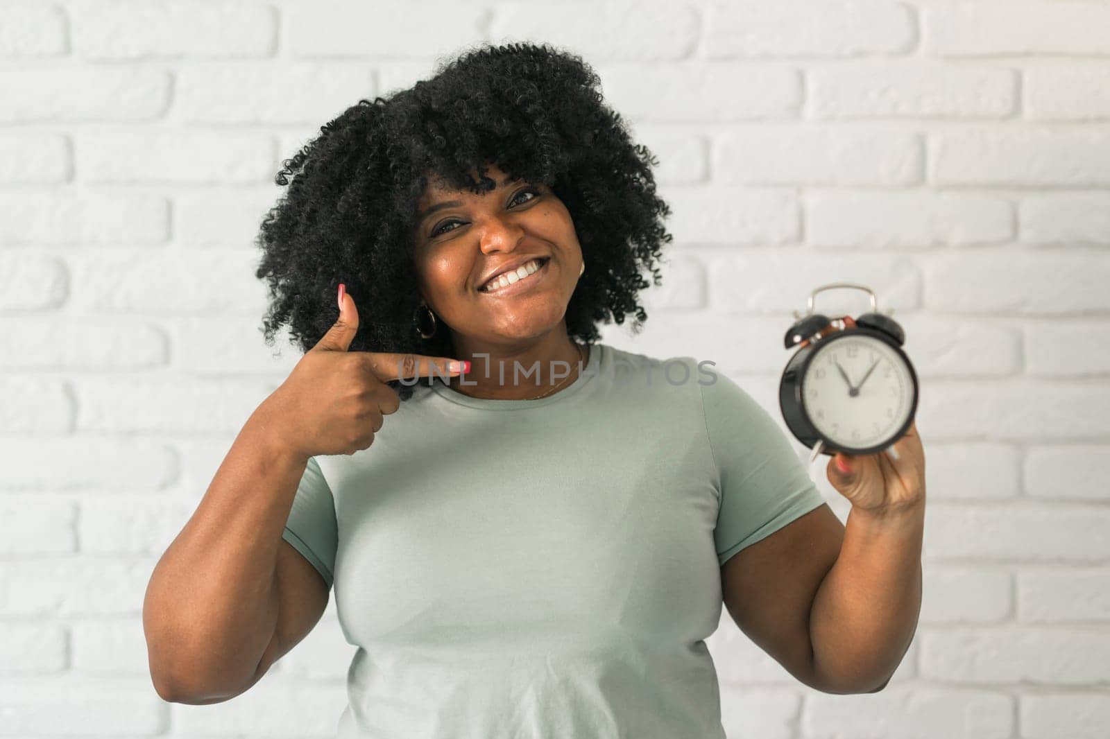 Surprised african american woman holding alarm clock amazed with open mouth and surprise happy face at home, on brick background. Being late and time passing quickly concept by Satura86