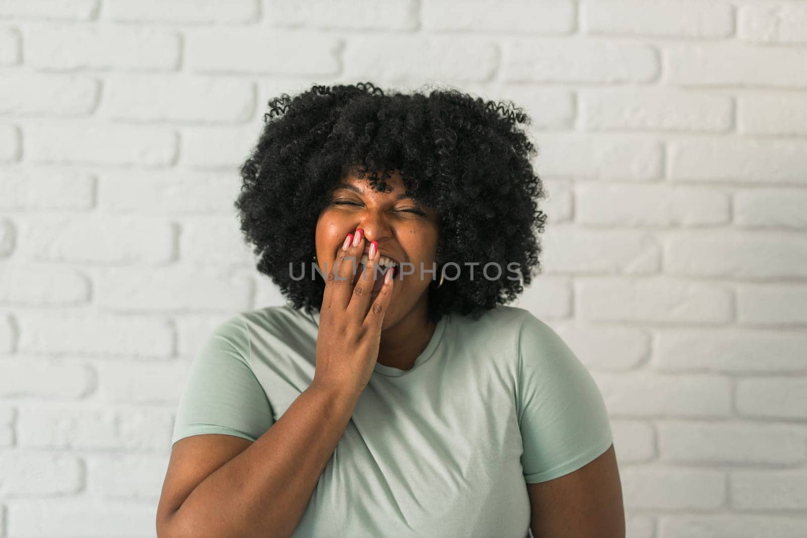 Laughing African American woman with an afro hairstyle and good sense of humor smiling and laugh on brick wall at home background. Happiness and good emotions