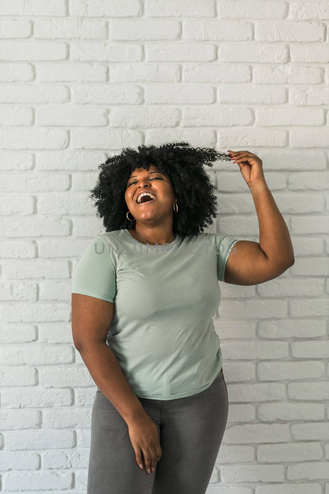 Laughing African American woman with an afro hairstyle and good sense of humor smiling and laugh on brick wall at home background. Copy space. Happiness and good emotions concept by Satura86