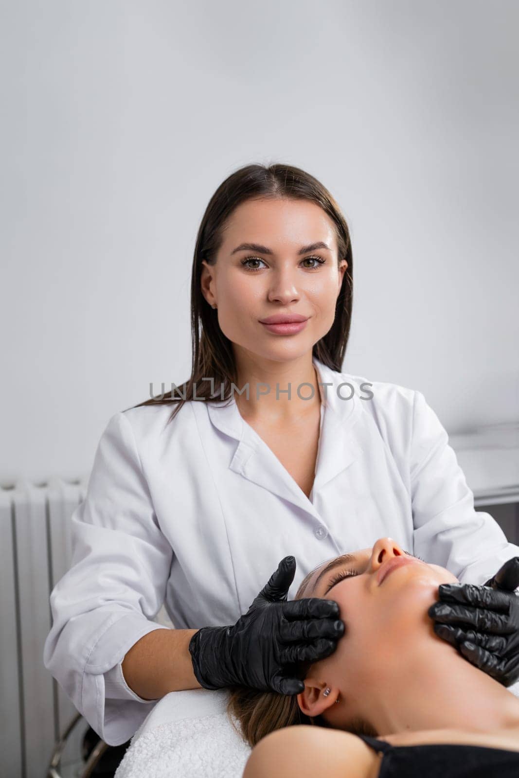 Portrait of a beautiful young woman cosmetologist does a facial lifting massage in a cosmetology clinic, vertical photo