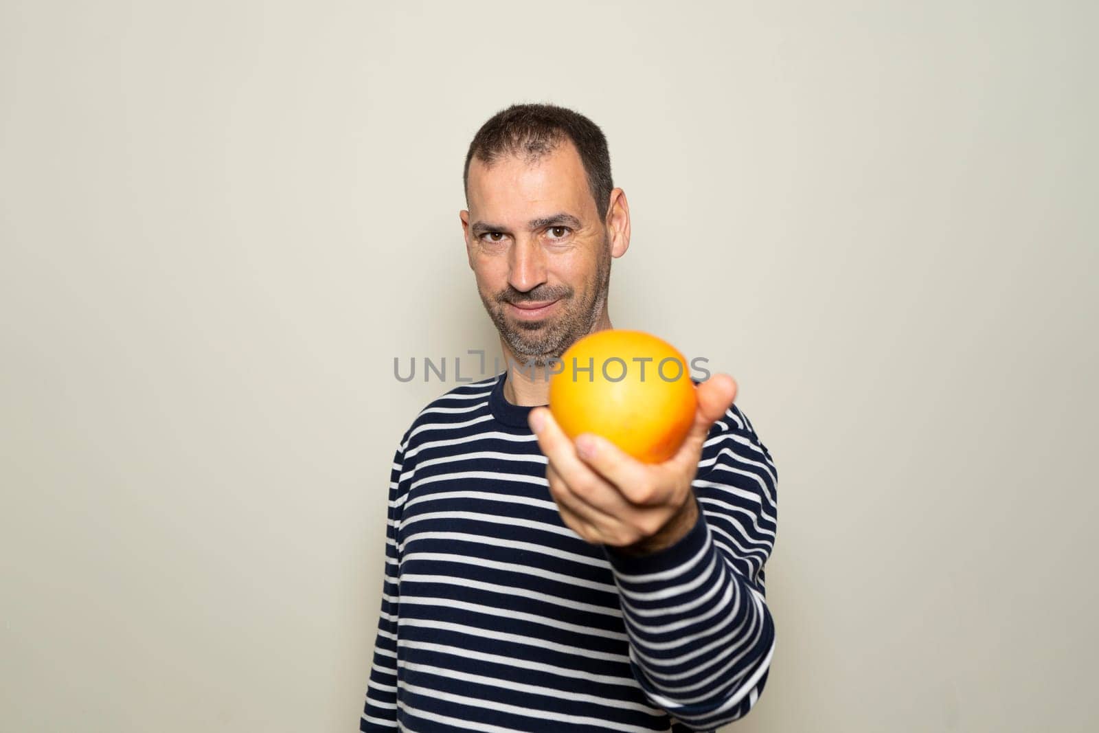 Bearded Hispanic man in his 40s wearing a striped sweater offering you an orange isolated on beige background. Healthy and recommended diet, Mediterranean diet, vitamin C. by Barriolo82